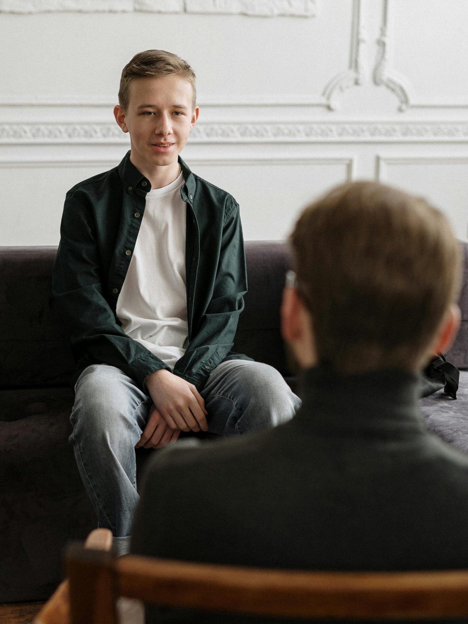 A teenage boy in a counseling session with a therapist in a modern loft setting, sitting on a sofa.