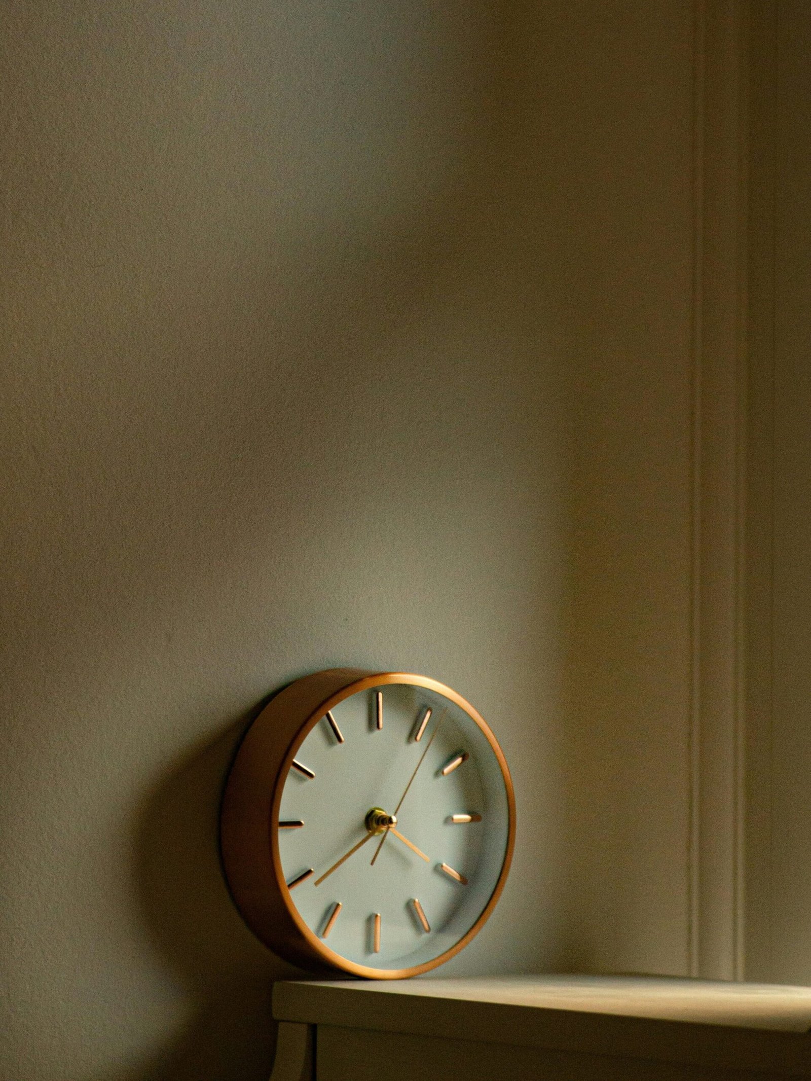 A retro-style analogue clock resting on a wood surface, casting warm light against a white wall.